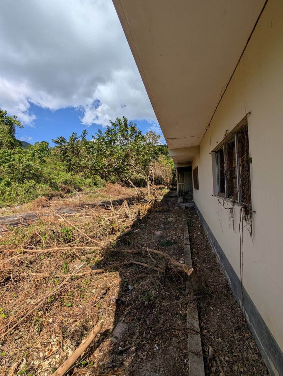 The side of a long single-storey building next to cleared land, with green hills behind